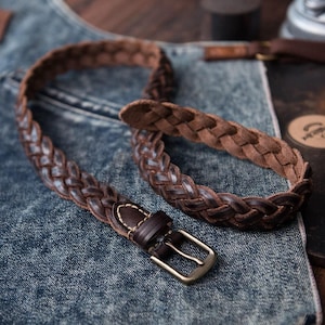 May include: A brown leather belt with a braided design. The belt features a brass buckle and a dark brown leather loop. The belt is lying on a denim surface.