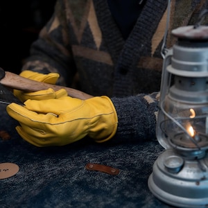 May include: Axe with a wooden handle and a metal head, held by a person wearing yellow leather gloves. A vintage lantern with a lit flame is in the background. The person is wearing a patterned sweater.