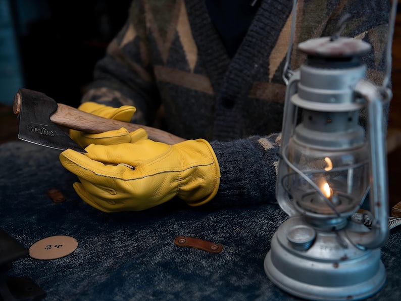 May include: Axe with a wooden handle and a metal head, held by a person wearing yellow leather gloves. A vintage lantern with a lit flame is in the background. The person is wearing a patterned sweater.