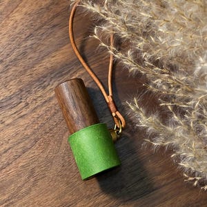 May include: A wooden and green leather accessory with a brown leather strap and gold-colored hardware. The cylindrical object has a dark brown wooden top and a bright green leather base. The strap is attached with a small gold-colored ring.