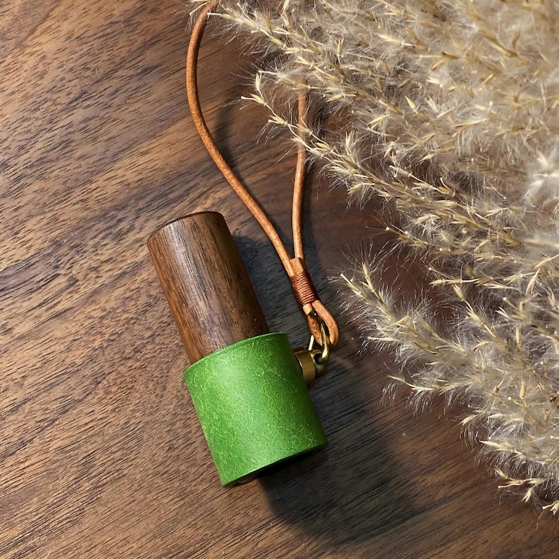 May include: A wooden and green leather accessory with a brown leather strap and gold-colored hardware. The cylindrical object has a dark brown wooden top and a bright green leather base. The strap is attached with a small gold-colored ring.