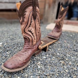 May include: A pair of brown leather cowboy boots with intricate stitching and an ostrich skin pattern. The boots are on a wooden boot stretcher.