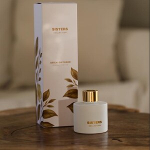 Könnte beinhalten: Ein weißer Stäbchendiffusor und eine passende Box aus der Sisters Collection. Der Diffusor hat einen goldenen Deckel und die Box zeigt goldene Blattmotive. Die Produkte werden auf einer Holzoberfläche präsentiert.