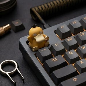 May include: A yellow Psyduck character artisan keycap sits on a mechanical keyboard with dark gray keys and gold lettering. The keyboard features a coiled cable, and a metal keycap puller is positioned nearby on the dark surface.