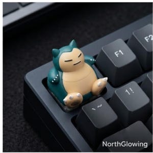 May include: A custom mechanical keyboard keycap shaped like a teal and beige Snorlax character with closed eyes, positioned on a black keyboard next to keys labeled F1 and 1!. The word NorthGlowing is printed in white at the bottom right corner.
