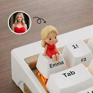 May include: A custom keyboard keycap featuring a small figurine of a blonde person wearing a red dress, seated on a white keycap labeled with the name Emma. The keycap is part of a white mechanical keyboard with visible key labels like 1!, F2, and Tab.
