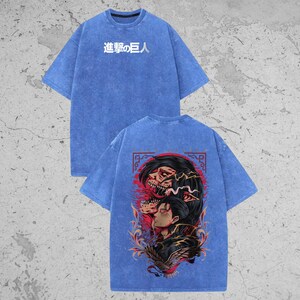 May include: A blue acid-wash t-shirt featuring the Japanese text "進撃の巨人" on the front chest. The back displays a detailed graphic illustration of a character with dark hair and a skeletal figure, accented by red and orange flame-like patterns.