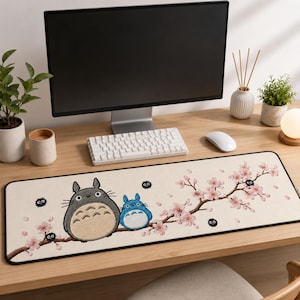 May include: A rectangular desk mat with a cream-colored surface featuring a brown tree branch with pink cherry blossoms, a large gray creature, a smaller blue creature, and several small black fuzzy soot sprites printed on the fabric.