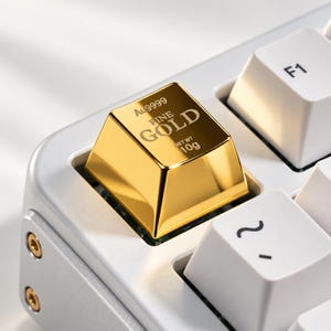 May include: A metallic gold-colored keyboard keycap shaped like a small ingot, featuring the engraved text Au9999, FINE GOLD, and NET WT 10g. The keycap is installed on a white mechanical keyboard next to a white F1 key and a tilde key.