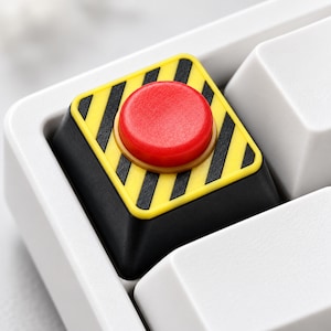 May include: A square mechanical keyboard keycap featuring a bright red circular button centered on a yellow and black diagonal hazard stripe pattern. The keycap has a black base and is positioned among several plain white keyboard keys.
