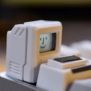 May include: A white mechanical keyboard keycap shaped like a vintage computer monitor with a small screen displaying a simple pixelated smiley face. The keycap features a molded plastic design with a separate keyboard component attached in front.