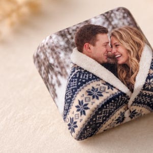 Può includere: Una calamita fotografica di forma quadrata con l'immagine stampata di due persone che si sorridono, avvolte in una coperta blu navy e color crema con un motivo a fiocchi di neve e una fodera in pile sherpa bianco sui bordi.