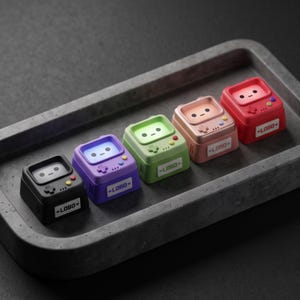 Peut inclure: Cinq touches de clavier mécanique carrées et petites, de couleurs noire, violette, verte, rose clair et rouge. Chaque touche présente un écran avec un visage simple, une croix directionnelle, deux boutons ronds et une étiquette blanche avec le texte LOBO.