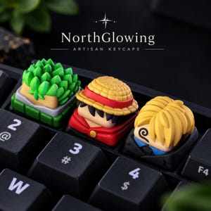 May include: Three custom artisan keycaps installed on a black mechanical keyboard. The left keycap features a green spiky plant design. The center keycap depicts a character with black hair and a yellow straw hat. The right keycap shows a blonde character.