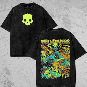 Könnte beinhalten: Schwarz gefärbtes T-Shirt mit einem neongrünen Totenkopf-Motiv auf der Vorderseite. Die Rückseite des Shirts zeigt eine farbenfrohe Grafik mit dem Wort "HELLDIVERS" und einem Totenkopf.