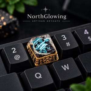 May include: A custom artisan keycap featuring a light blue dragon figure encased in clear resin, set atop a gold-colored base with engraved hieroglyphic-style symbols. The keycap is installed on a black mechanical keyboard between the 2 and 3 keys.
