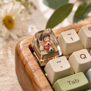 May include: A custom mechanical keyboard keycap featuring a clear resin housing that encapsulates a miniature figurine of a girl with brown hair wearing a pink outfit, sitting on a tatami mat with a small white creature and rice balls.