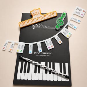 Könnte beinhalten: Ein schwarzes Musiknotizbuch mit einem Klaviertasten-Design, ein Stift mit einem Klaviertastenmuster und verschiedene Musik-Accessoires. Enthält einen Violinschlüssel, einen Clip in Klaviertastenform und Notenbezeichnungen.
