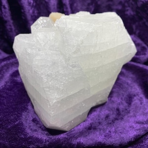 May include: A large, translucent white selenite crystal cluster. The crystal has a layered appearance with a few small, light-colored inclusions. The selenite is displayed against a deep purple velvet background.