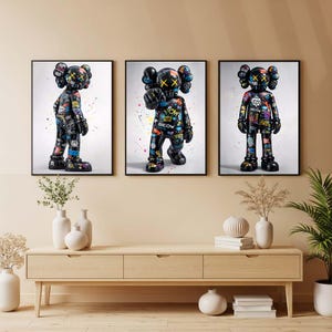 Puede incluir: Tres impresiones artísticas enmarcadas que presentan una figura negra con ojos en forma de cruz y un diseño de graffiti colorido. La figura se representa en varias poses sobre un fondo blanco, con marcos negros. La obra de arte se exhibe sobre un mueble de madera.