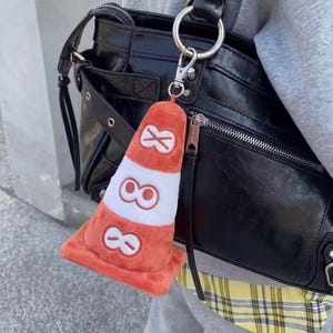 Puede incluir: Un llavero de peluche con forma de cono de tráfico naranja y blanco, sujeto a un bolso de cuero negro. El cono presenta tres conjuntos de gráficos blancos y naranjas. El bolso tiene herrajes plateados y una cremallera.