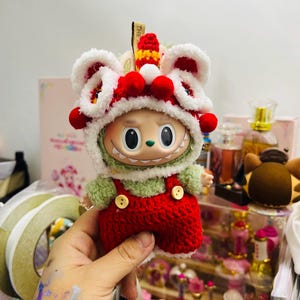 May include: A whimsical doll wearing a red and white lion dance hat with red pom-poms. The doll has a green fuzzy shirt and red overalls with yellow buttons. The doll is being held in a hand.
