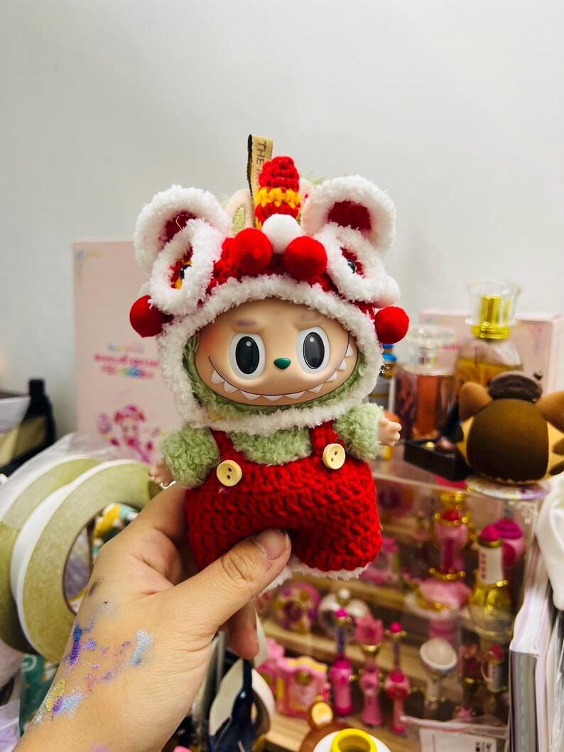 May include: A whimsical doll wearing a red and white lion dance hat with red pom-poms. The doll has a green fuzzy shirt and red overalls with yellow buttons. The doll is being held in a hand.