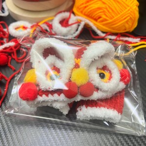 May include: A small, handmade lion dance costume in a clear plastic bag. The costume is primarily red and white, with yellow and red accents. The lion's face has large, expressive eyes and a smiling mouth. Red yarn and yellow yarn are in the background.