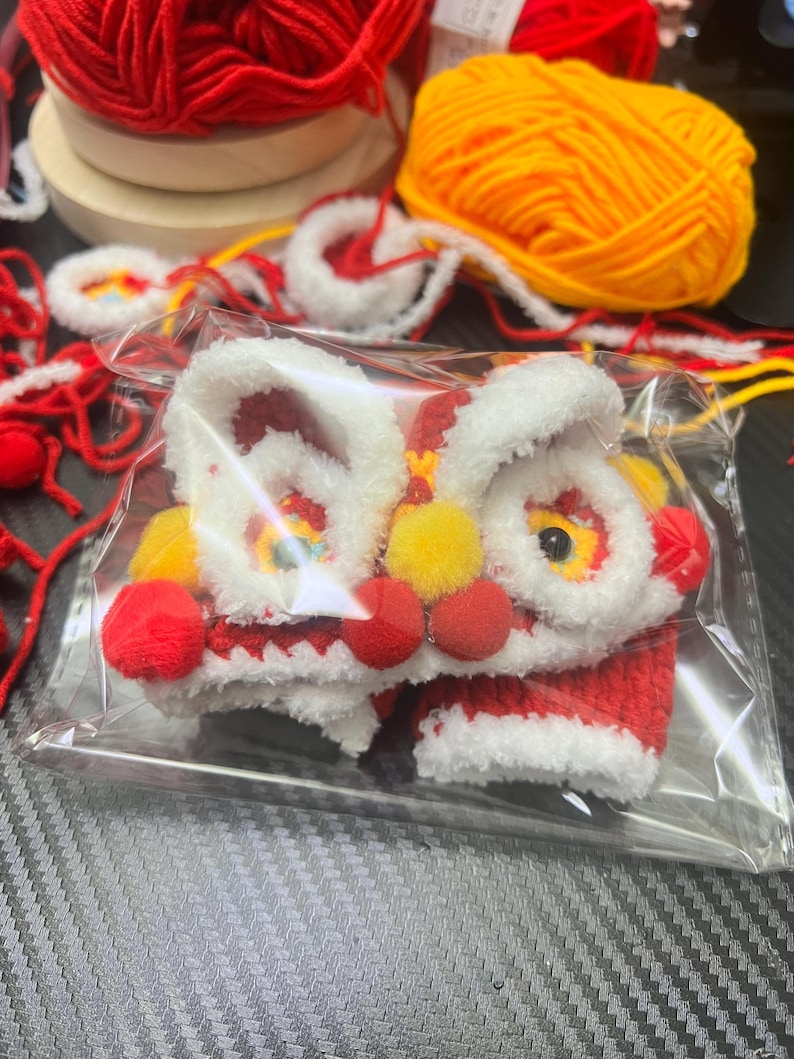 May include: A small, handmade lion dance costume in a clear plastic bag. The costume is primarily red and white, with yellow and red accents. The lion's face has large, expressive eyes and a smiling mouth. Red yarn and yellow yarn are in the background.