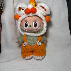 May include: A whimsical doll wearing a white and orange crocheted hat with orange pom-poms and a horn. The doll has a green fuzzy shirt and orange overalls with white trim. The doll has a smiling face with large black eyes.