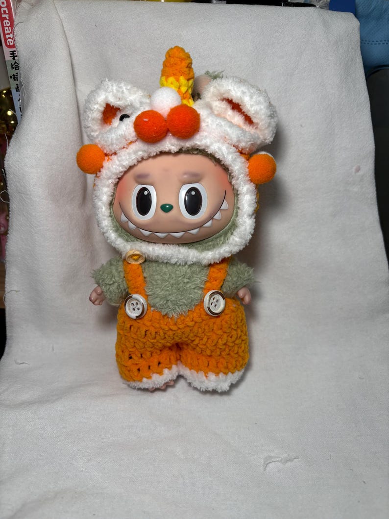 May include: A whimsical doll wearing a white and orange crocheted hat with orange pom-poms and a horn. The doll has a green fuzzy shirt and orange overalls with white trim. The doll has a smiling face with large black eyes.