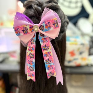 May include: A large, colorful hair bow featuring a central jewel and multiple layers of pink, purple, and blue ribbons. The bow has a mesh overlay and long, dangling ribbons with a repeating cartoon character pattern. The bow is attached to dark hair.
