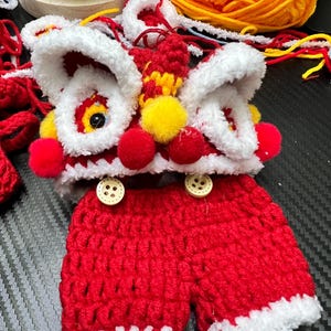 May include: A handmade crochet outfit in the style of a Chinese lion dance costume. The outfit includes a red and white lion headpiece with yellow and red accents, and red shorts with white trim. Two wooden buttons are on the shorts.