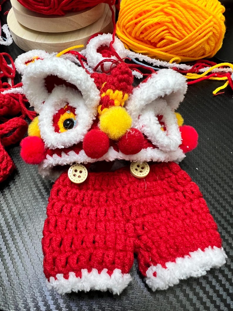 May include: A handmade crochet outfit in the style of a Chinese lion dance costume. The outfit includes a red and white lion headpiece with yellow and red accents, and red shorts with white trim. Two wooden buttons are on the shorts.