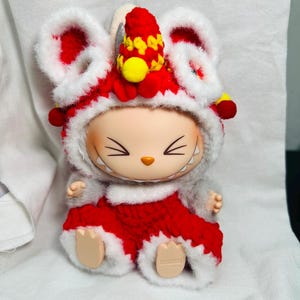 May include: A collectible figurine of a mouse wearing a red and white lion dance costume. The costume features large ears, a red and yellow headdress, and a red outfit with white trim. The figurine has a smiling face and is sitting down.