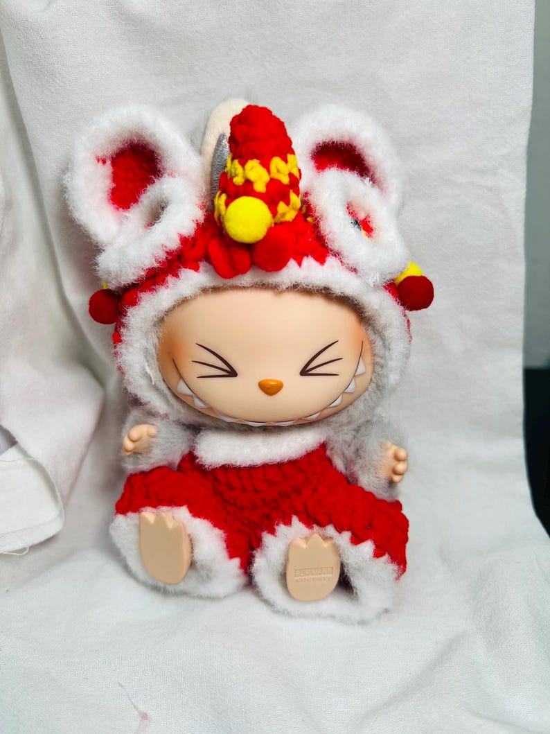 May include: A collectible figurine of a mouse wearing a red and white lion dance costume. The costume features large ears, a red and yellow headdress, and a red outfit with white trim. The figurine has a smiling face and is sitting down.