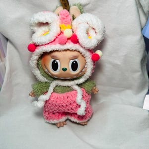 May include: A whimsical doll wearing a handmade pink and white crochet outfit with a matching hood. The hood is adorned with white fluffy trim, pink pom-poms, and small yellow accents. The doll has large, expressive eyes and a cheerful expression.