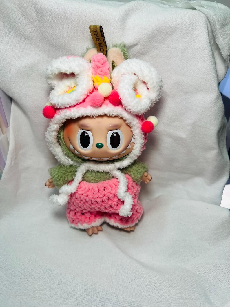 May include: A whimsical doll wearing a handmade pink and white crochet outfit with a matching hood. The hood is adorned with white fluffy trim, pink pom-poms, and small yellow accents. The doll has large, expressive eyes and a cheerful expression.