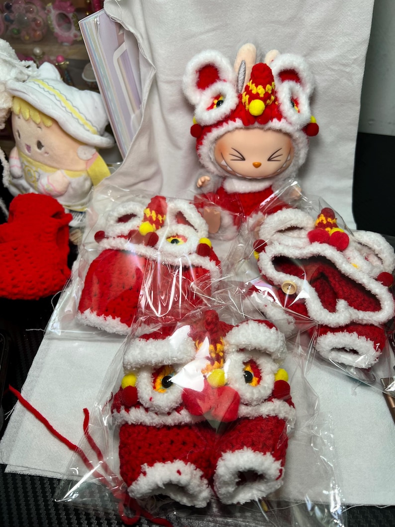 May include: A collection of red and white crocheted lion dance costumes and a doll wearing a matching outfit. The costumes feature intricate details, including yellow accents and fluffy white trim. The doll has a rabbit-like face.