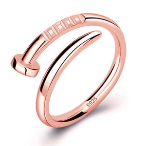 Peut inclure: Bague de couleur or rose en forme de clou. La bague présente une bande incurvée qui s'enroule autour du doigt, avec une tête de clou et des détails texturés. La bague est marquée "S925".