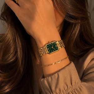 May include: A gold-tone wristwatch with a rectangular case and a deep green dial featuring gold-tone hands and hour markers. The watch has a multi-link metal bracelet and is accompanied by a thin gold-tone chain bracelet on the same wrist.