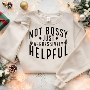 Könnte beinhalten: Ein cremefarbenes Sweatshirt mit dem Text "NOT BOSSY JUST AGGRESSIVELY HELPFUL" in Schwarz. Das Design beinhaltet einen Stern und eine Hand, die ein Friedenszeichen mit einer Skeletthand macht. Das Sweatshirt ist von Weihnachtsdekorationen umgeben.