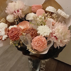 Peut inclure: Un bouquet de fleurs de savon aux tons rose, pêche et blanc, agrémenté de verdure et de tulle. Le bouquet comprend des pivoines, des roses et d'autres formes florales, enveloppé dans du papier brun et présenté dans un vase en verre transparent.