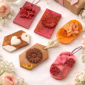 Peut inclure: Assortiment de fondants parfumés décoratifs de différentes formes et couleurs, notamment des étiquettes rectangulaires et ovales. Chaque étiquette présente des ornements floraux ou en forme de cœur. La palette de couleurs comprend des nuances de rose, d'orange et de marron, avec une boîte cadeau et une bougie en arrière-plan.