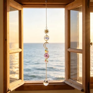 Op de afbeelding: Een hangend zonnevanger-ornament met een goudkleurige ketting, een zon-bedel, een ronde hanger met blauwe gedroogde bloemen, een maansikkel, twee iriserende glazen bollen, een paarse bloemschijf en een heldere kristallen druppelvormige hanger.
