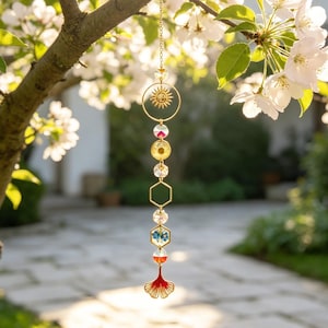 Op de afbeelding: Een hangende zonnevanger met een goudkleurige metalen ketting, een zon-bedel, heldere gefacetteerde kralen, twee zeshoekige frames met gedroogde bloemen en een rode, ginkgobladvormige metalen hanger aan de onderkant.