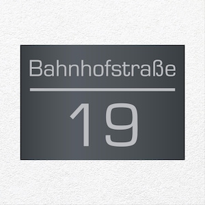 Puede incluir: Placa rectangular de dirección con el texto "Bahnhofstraße" y el número "19" en gris claro. El fondo es gris oscuro. La placa está montada en una pared blanca texturizada.