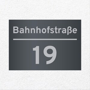 Könnte beinhalten: Ein rechteckiges, dunkelgraues Hausnummernschild mit dem Text Bahnhofstraße in hellgrauer serifenloser Schrift über einer horizontalen Linie, wobei die Zahl 19 in einer großen, fetten hellgrauen Schrift unter der Linie zentriert ist.