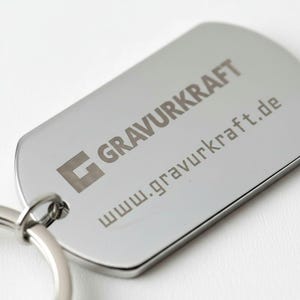 Könnte beinhalten: Ein silberfarbener Schlüsselanhänger in Form einer Hundemarke mit dem eingravierten Text "GRAVURKRAFT" und "www.gravurkraft.de". Der Schlüsselanhänger ist an einem silbernen Ring befestigt.