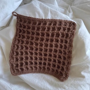 Puede incluir: Un paño de lavado cuadrado de ganchillo marrón con una textura similar a un gofre. El paño de lavado tiene un pequeño lazo para colgar y está hecho de un hilo suave y texturizado. Se muestra sobre una superficie blanca arrugada.